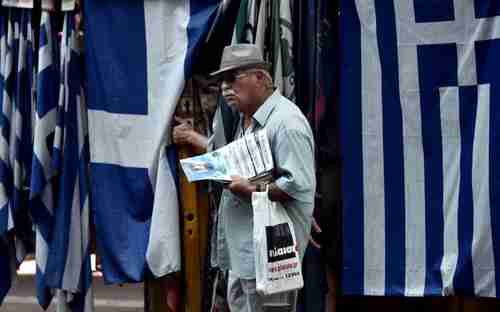 An old man carrying newspapers stands in front of a row of Greek flags in Athens (Kathimerini)